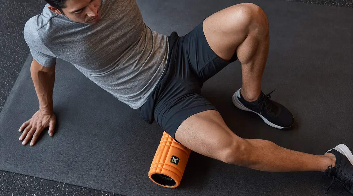 Close-up of hands placing a TriggerPoint GRID foam roller under the back of a thigh on a gym floor